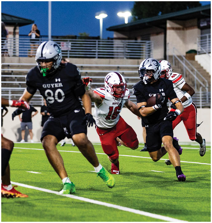 Adults in football uniforms are actively running on a green field during a game at dusk with stadium seating in the background. Description generated by AI