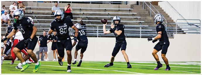 People in football uniforms are on a field with one person preparing to throw a football while others move around, and spectators are seated in the background. Description generated by AI