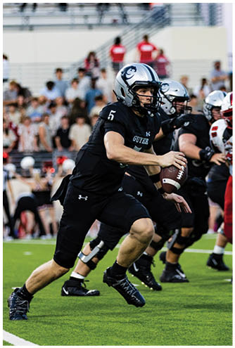 People wearing red shirts stand in the background of a stadium while a person in the foreground wears a silver football helmet with a panther logo. Description generated by AI