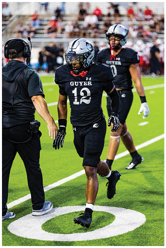 Two people wearing black football uniforms and helmets are on a field with blurred spectators in the background. Description generated by AI