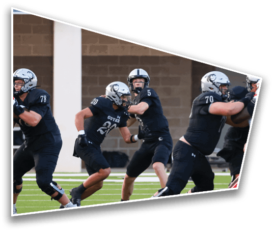 Several people in football uniforms are engaged in a play on a green field with a brick wall background. Description generated by AI