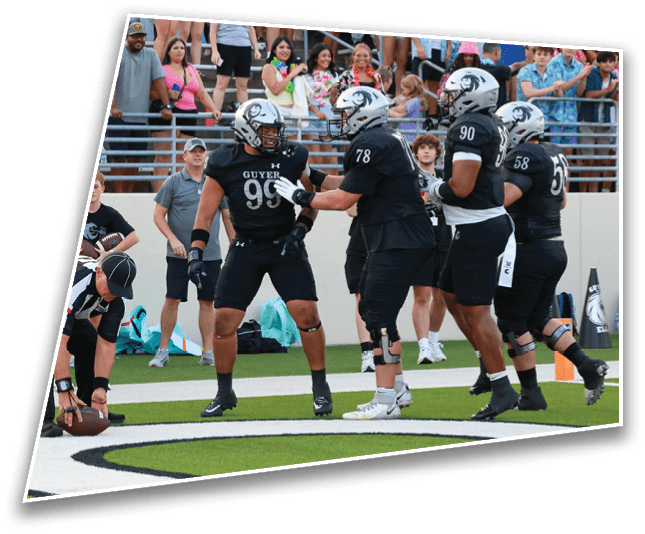 A group of football players in black uniforms and helmets are gathered near the end zone while a referee places the football on the ground, with a crowd of people watching from the stands. Description generated by AI