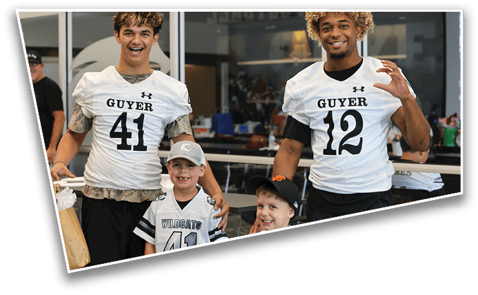Two adults wearing football jerseys with the numbers 41 and 12 stand behind two children wearing sports apparel, all smiling indoors. Description generated by AI