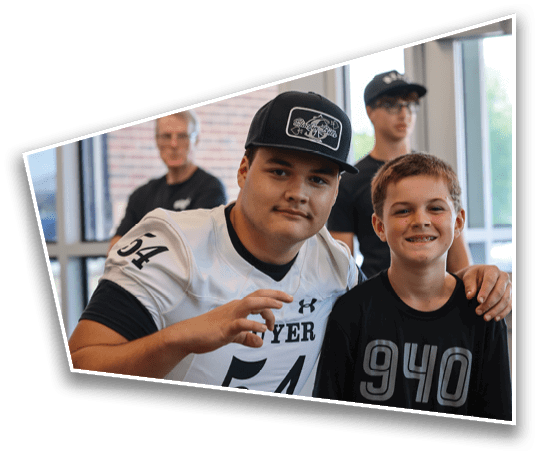 Two people are sitting closely together indoors with one wearing a sports jersey and cap while smiling, and two others are visible in the background near large windows. Description generated by AI