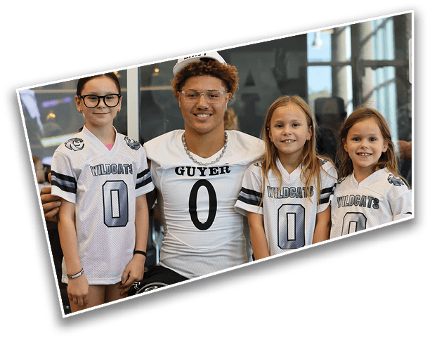 An adult wearing a sports jersey with the number zero is seated with three children standing beside them, all wearing matching sports jerseys with the number zero and smiling indoors. Description generated by AI