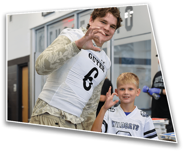 A person wearing a sports jersey with the word “GUYER" is making a hand gesture and smiling indoors near a wall with framed sports memorabilia. Description generated by AI