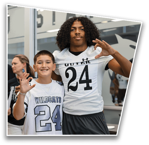 Two people are smiling indoors, with one wearing a sports jersey and making a hand gesture. Description generated by AI