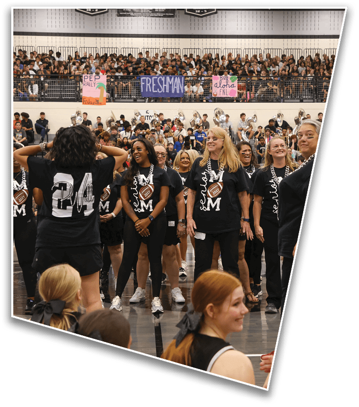 People wearing matching black shirts with “Senior" and "M" printed on them stand in a gymnasium filled with a large crowd and colorful banners reading "Pep Rally," "Freshman," and "Aloha." Description generated by AI