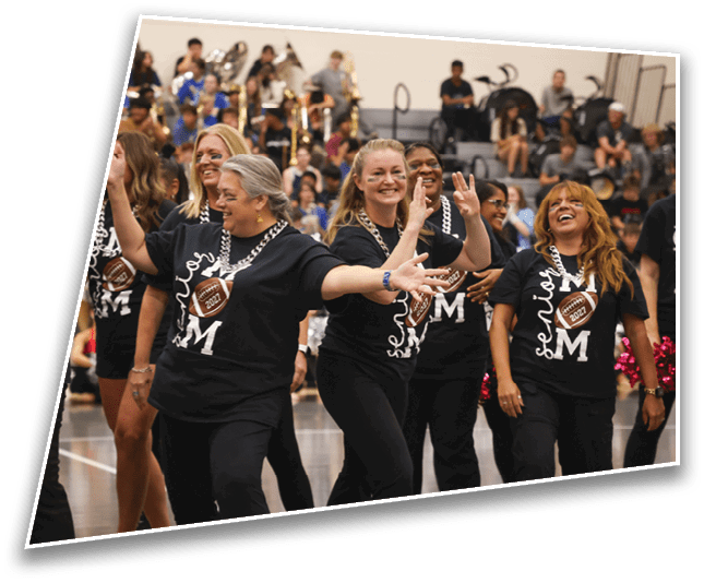 A group of adults wearing matching black shirts with a football design are smiling and dancing in a gymnasium with a band playing in the background. Description generated by AI