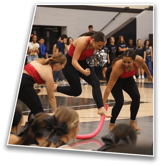 Two adults wearing red tops and black pants are performing a lively gym routine involving a pink foam noodle while a group of people watch in the background. Description generated by AI