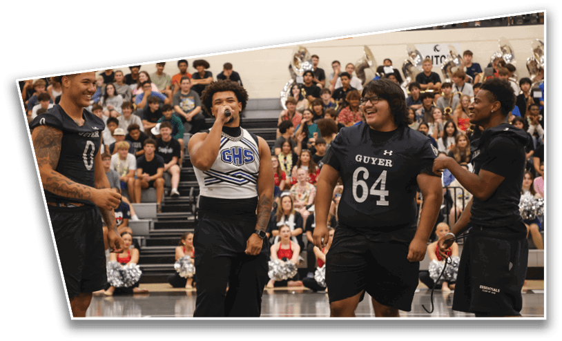 Four people in athletic clothing stand on a gymnasium floor with a crowd seated in bleachers behind them, one person holding a microphone and another wearing a shirt with “GUYER 64" printed on it. Description generated by AI