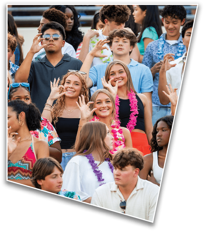 A group of smiling people in casual clothing are gathered closely together, some making hand gestures, with one person wearing a pink lei. Description generated by AI