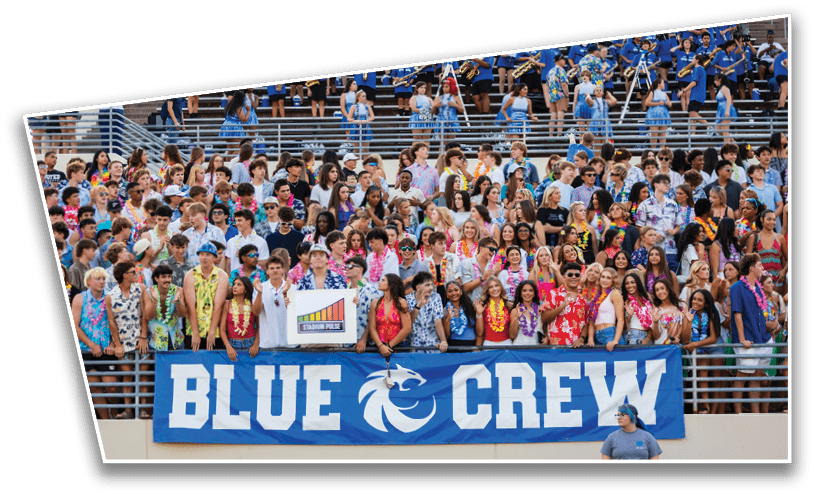 A large group of people wearing colorful Hawaiian shirts and leis are gathered in stadium bleachers behind a blue banner that reads “BLUE CREW" with a wildcat logo. Description generated by AI