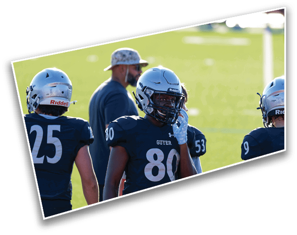 People wearing football helmets and uniforms stand on a field with one person adjusting their helmet while another person in casual clothing and a hat stands nearby. Description generated by AI
