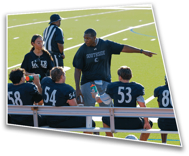 A coach wearing a Southside shirt is pointing and talking to four seated football players in uniform on a bench while a referee and another person stand nearby on a sports field. Description generated by AI
