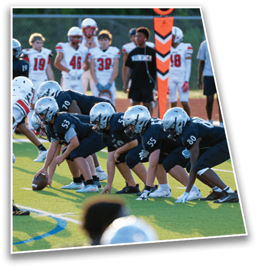 Children in football uniforms are lined up on a field facing each other, preparing for a play during a game with other players and a coach standing in the background. Description generated by AI