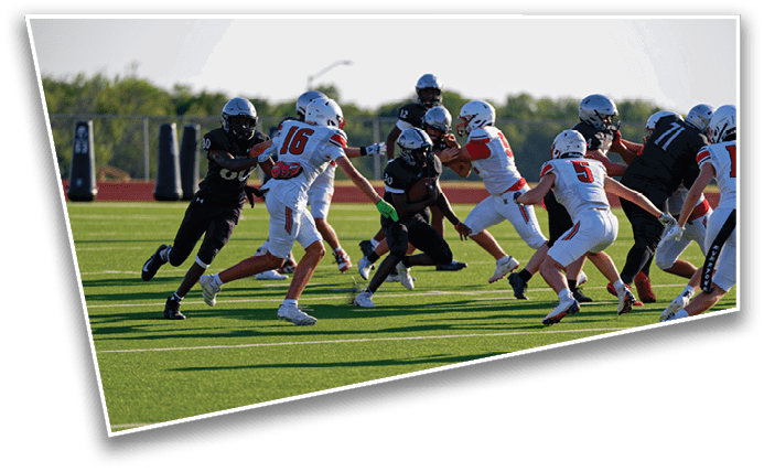 People wearing helmets and sports uniforms are actively playing football on a green field with trees in the background. Description generated by AI