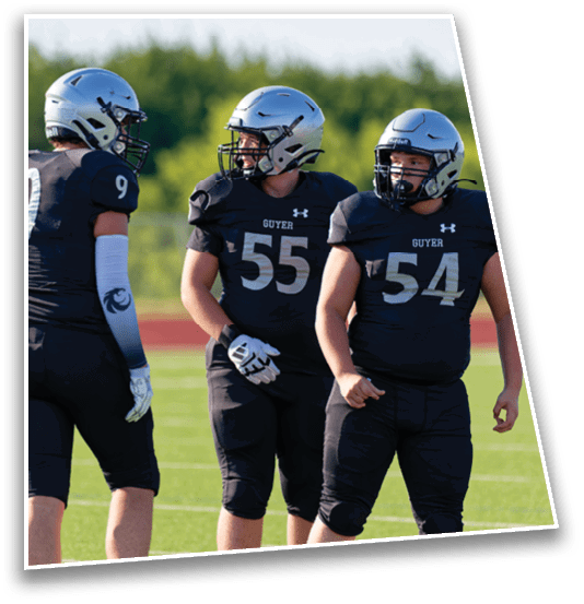 Three people in football uniforms and helmets stand on a grassy field with a track and trees in the background. Description generated by AI