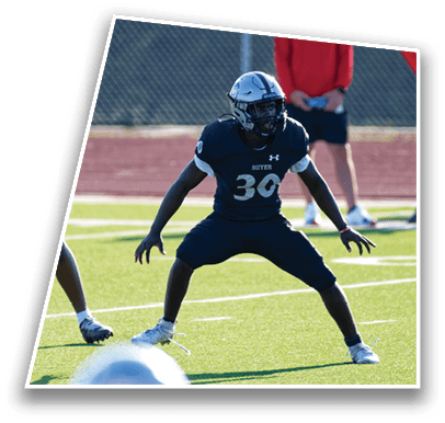 A person wearing a dark sports uniform with the number 30 is in a defensive stance on a football field with another person partially visible in the foreground. Description generated by AI