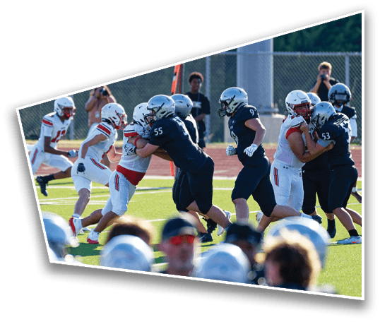 People in football uniforms are engaged in blocking and tackling on a sunlit field during a game. Description generated by AI