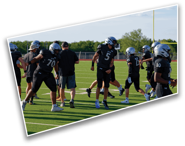 People wearing black football uniforms and helmets are gathered on a green football field near a goalpost under a clear sky. Description generated by AI