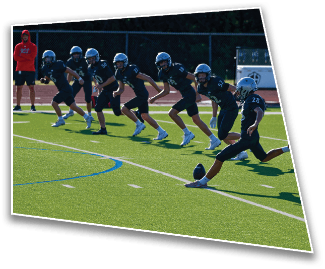 A group of people in football uniforms and helmets are lined up on a field with one person kicking a football. Description generated by AI