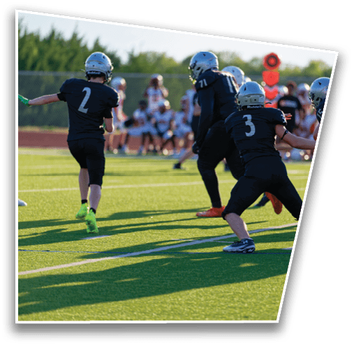 People wearing helmets and sports uniforms are playing football on a grassy field with one person preparing to throw the ball while others are positioned to block or receive. Description generated by AI