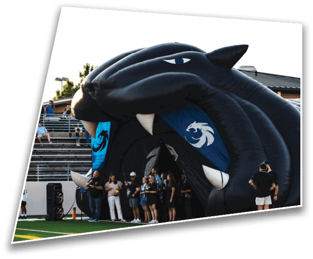 A group of people stands inside and around a large inflatable black panther head on a sports field with bleachers in the background. Description generated by AI