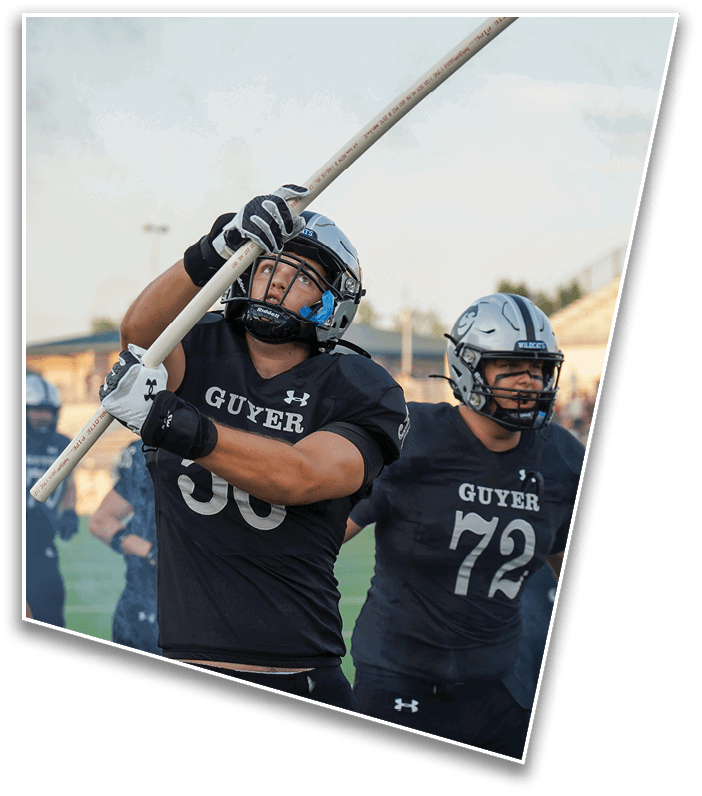 Two people wearing football helmets and black jerseys with the word “GUYER" are on a field, with one holding a pole and the other standing behind. Description generated by AI