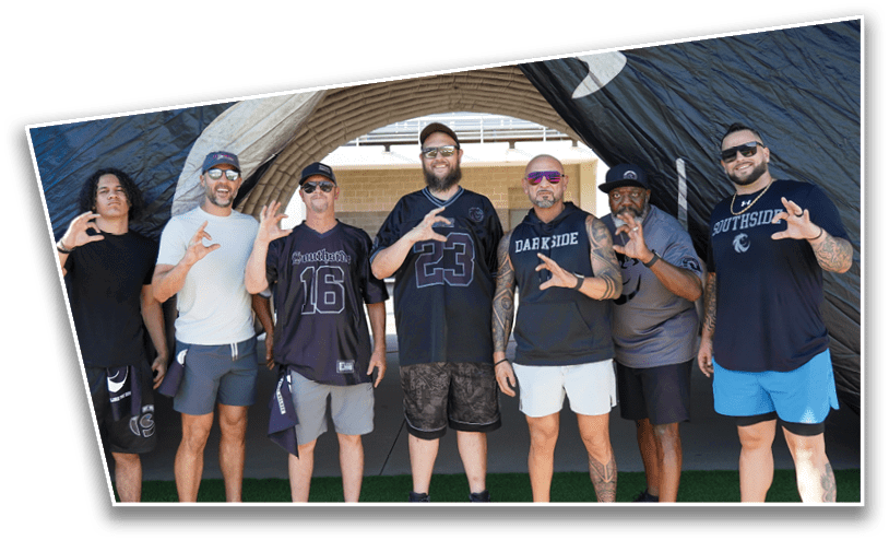 Seven adults wearing casual sportswear stand on grass under a curved structure, each making a hand gesture with three fingers extended. Description generated by AI