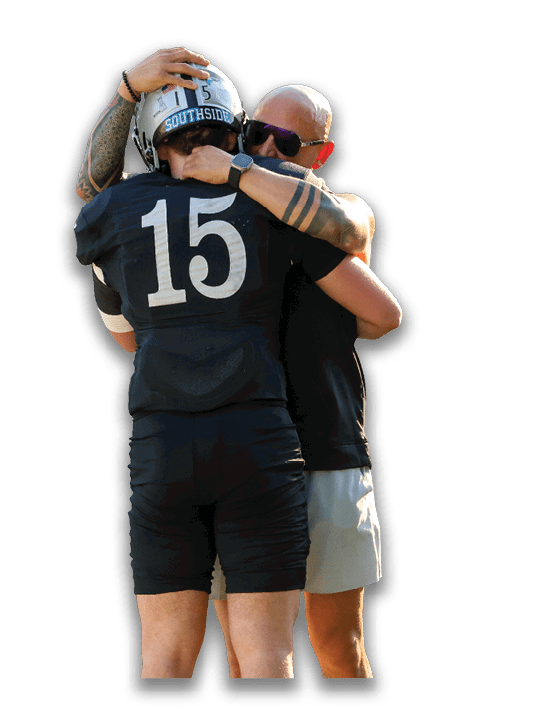 An adult wearing sunglasses and a smartwatch embraces a person in a football uniform with the number 15 and a helmet labeled Southside. Description generated by AI