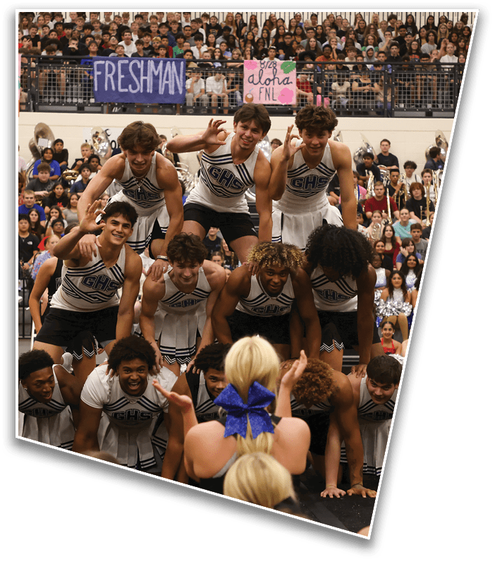 A group of people in cheerleading uniforms form a human pyramid in front of a large crowd in a gymnasium with banners reading “FRESHMAN" and "PEP RALLY." Description generated by AI