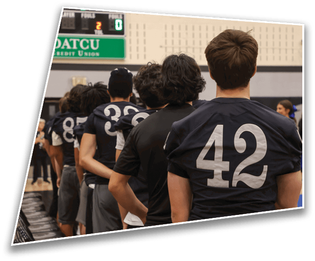A group of people is watching a basketball game in a gymnasium with a scoreboard showing the visitor team leading and an American flag hanging on the wall. Description generated by AI