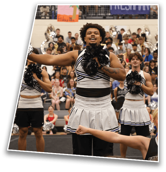 A group of adults in cheerleading uniforms perform with pom-poms in a gymnasium with a crowd and band in the background. Description generated by AI