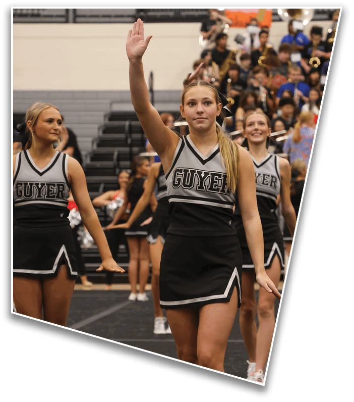 A person in a cheerleading uniform with the word “GUYS" raises one hand while standing in a gymnasium with other people and bleachers in the background. Description generated by AI
