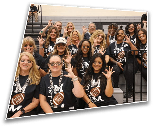 A group of adults wearing matching black shirts with a football design and face paint are sitting on bleachers smiling and making a hand gesture. Description generated by AI