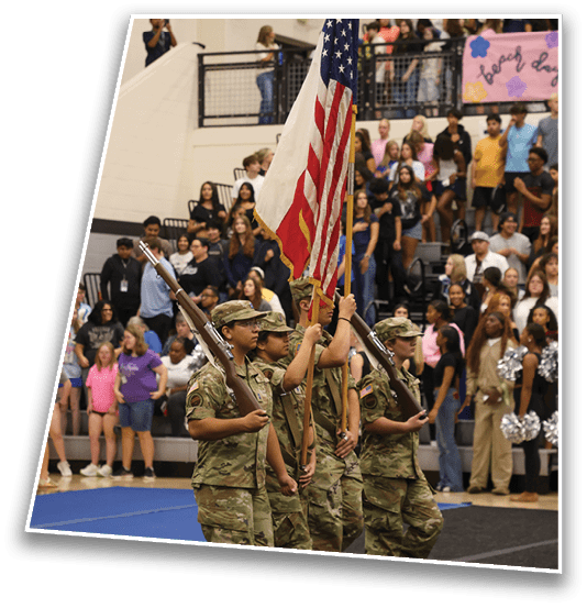 An American flag is displayed indoors in front of a crowd of people seated and standing on bleachers in a gymnasium or auditorium. Description generated by AI