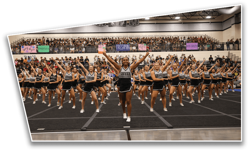 A large group of people in matching cheerleading uniforms perform a synchronized routine on a gym floor with a crowd watching from bleachers behind them. Description generated by AI