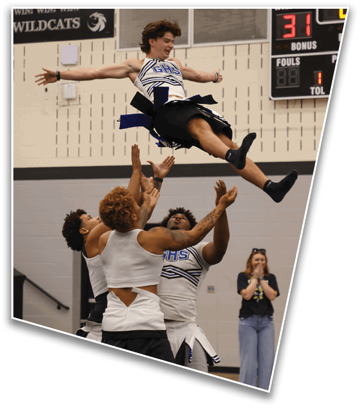 A person wearing a cheerleading uniform is being lifted into the air by several hands during a cheerleading stunt inside a gymnasium. Description generated by AI