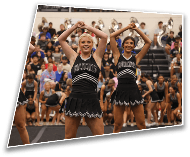 Two adults in black cheerleading uniforms with “Wildcats" on the front are performing a routine with their arms raised above their heads in a gymnasium filled with spectators and a band in the background. Description generated by AI