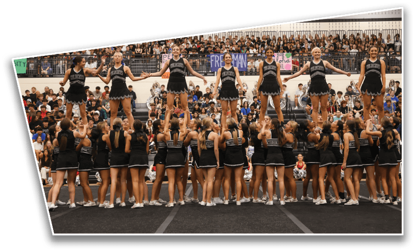 A large group of people in matching black cheerleading uniforms perform a coordinated stunt in a gymnasium filled with seated spectators. Description generated by AI