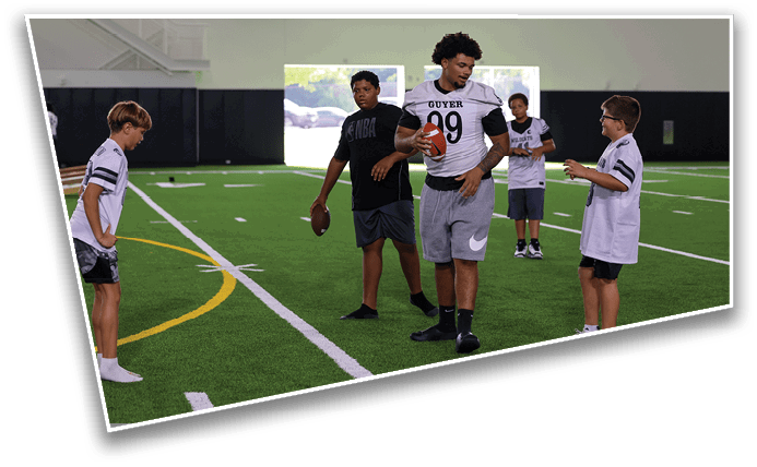 Five people wearing sports attire stand on an indoor football field, with one person holding a football and appearing to instruct or demonstrate to the others. Description generated by AI