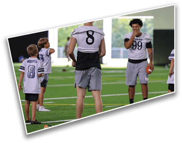 Several people wearing sports jerseys with numbers are standing on an indoor turf field, with one person holding a football. Description generated by AI