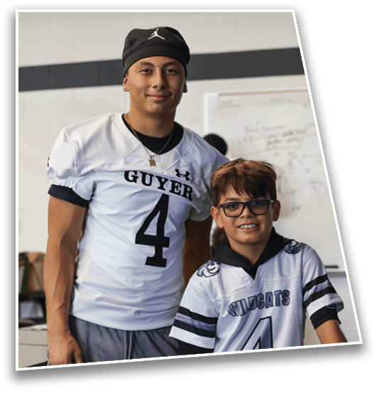Two people wearing football jerseys with the number 4 stand indoors smiling, one in a white Guyer jersey and the other in a white Wildcats jersey with glasses. Description generated by AI