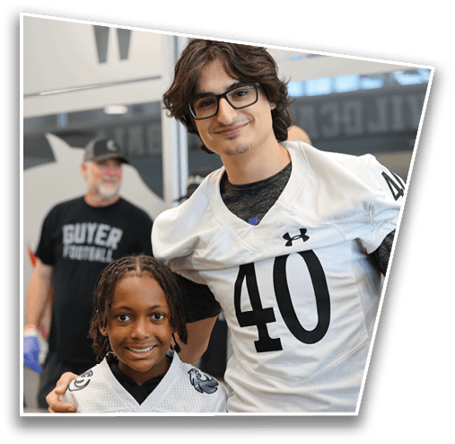 A person with medium-length wavy hair and glasses is smiling while wearing a white sports jersey over a dark shirt in an indoor setting. Description generated by AI