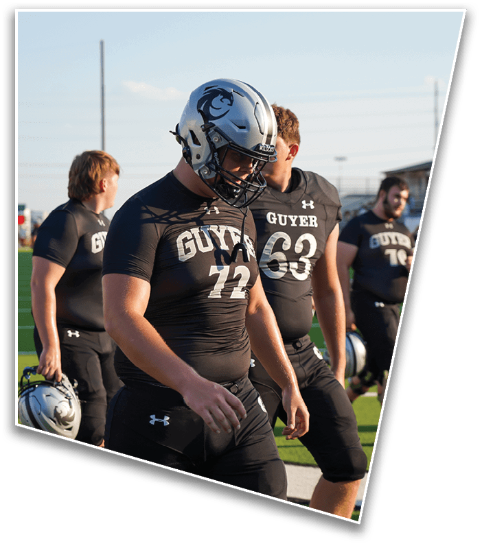 Several people wearing black football uniforms with “GUYER" printed on them are standing on a sports field, with one person wearing a silver helmet and others in the background. Description generated by AI
