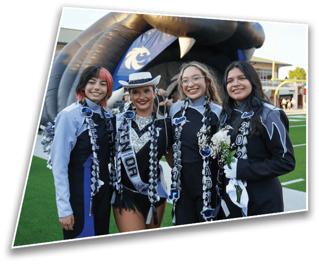 Four people wearing band uniforms and sashes are standing together smiling on a football field with an inflatable mascot in the background. Description generated by AI