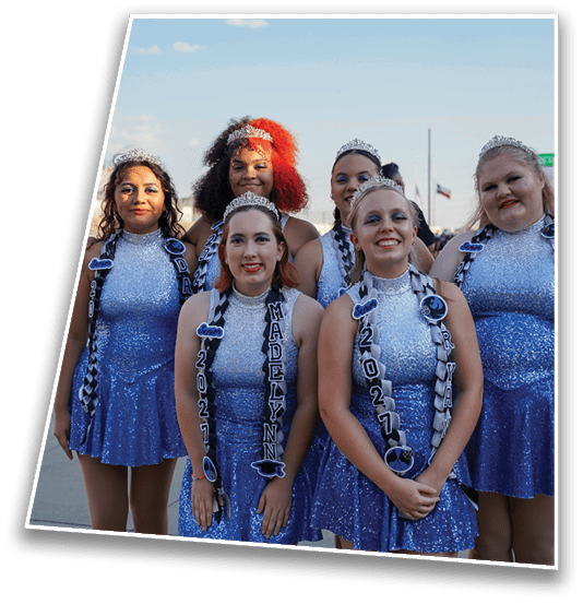 Five people wearing sparkly blue dresses and tiaras stand together outdoors, smiling and posing for a photo with sashes displaying names and years. Description generated by AI