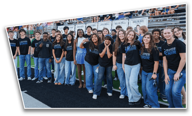 A group of people wearing matching choir t-shirts stand together on a sports field with bleachers and posters in the background. Description generated by AI