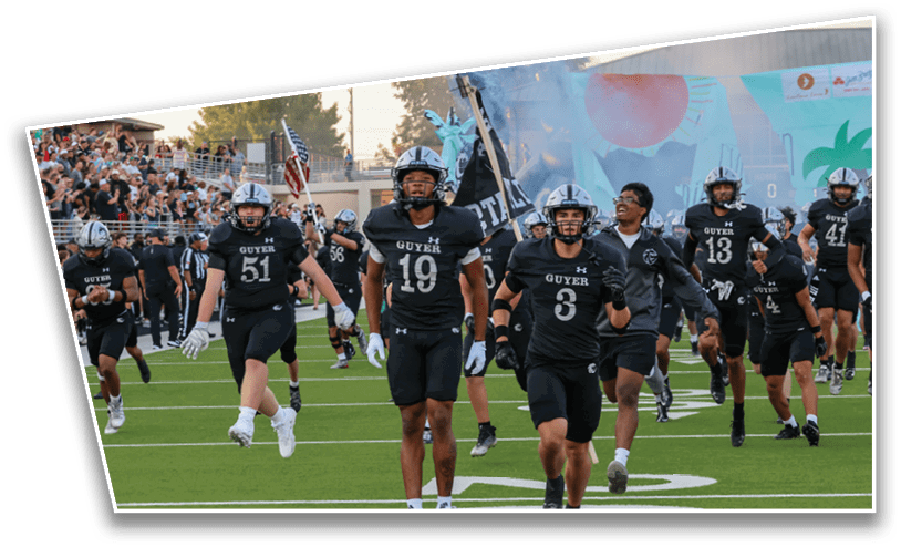 A group of people in black sports uniforms with helmets are running onto a football field with flags and smoke in the background, while a crowd watches from the stands. Description generated by AI